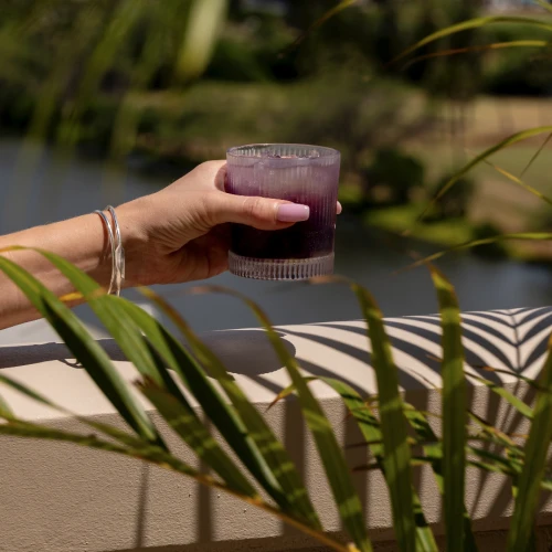 A hand holds a purple drink in a glass against a backdrop of greenery and buildings, viewed through palm leaves on a sunny day.
