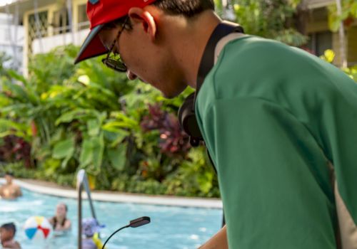 A poolside DJ mixes tracks on turntables, wearing a red cap and green shirt as swimmers relax in the background.