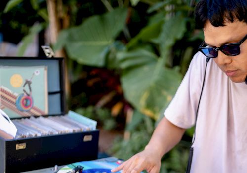 A DJ is spinning colorful vinyl records outdoors, with equipment and a record case nearby. They are wearing sunglasses.
