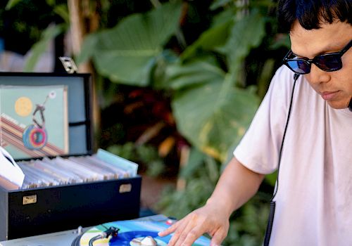 A DJ is spinning colorful vinyl records outdoors, with equipment and a record case nearby. They are wearing sunglasses.