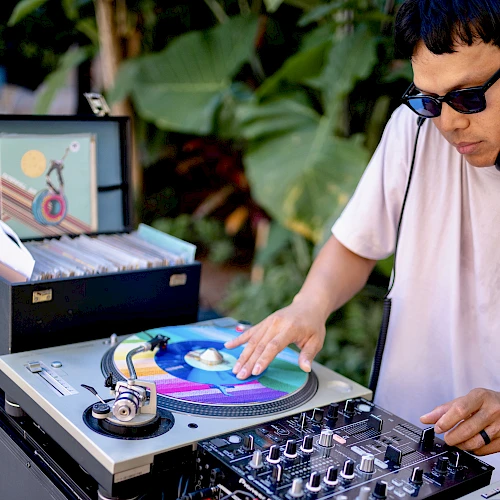 A DJ is spinning colorful vinyl records outdoors, with equipment and a record case nearby. They are wearing sunglasses.