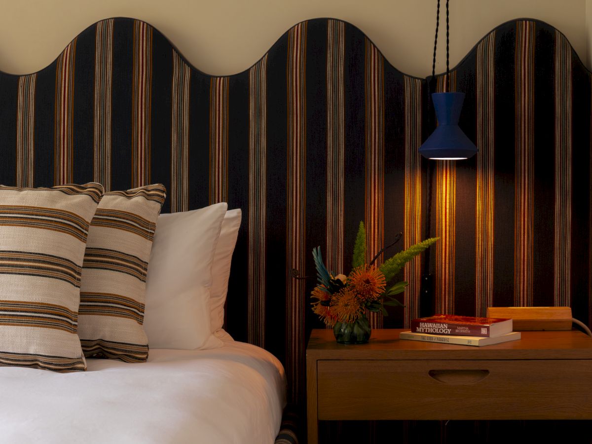 Ein gem&uuml;tliches Schlafzimmer mit geschwungenem Kopfteil, gestreiften Kissen, einem Nachttisch mit Lampe, B&uuml;chern und Blumen, die eine warme Atmosph&auml;re schaffen.