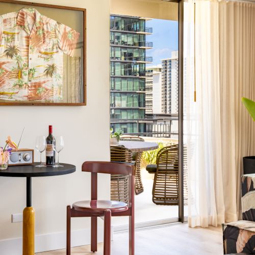 Stilvolles Zimmer mit gerahmtem Hemd, zwei St&uuml;hlen, Wein auf dem Tisch, Sofa mit gemusterten Kissen und Balkon mit Blick auf die Stadtgeb&auml;ude.