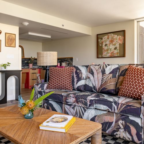 Ein Wohnzimmer mit einem Sofa mit Blumenmuster, einem Holztisch mit B&uuml;chern und gepunkteten Kissen, dekoriert mit gerahmten Bildern und Pflanzen.