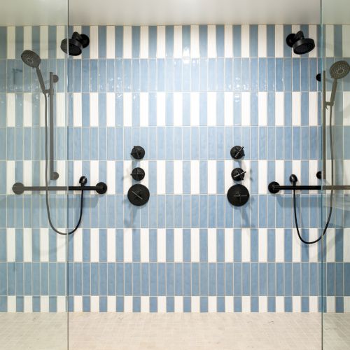 Das Bild zeigt eine moderne Dusche mit zwei Duschk&ouml;pfen, blau-wei&szlig; gestreiften Fliesen und schwarzen Armaturen an den W&auml;nden.