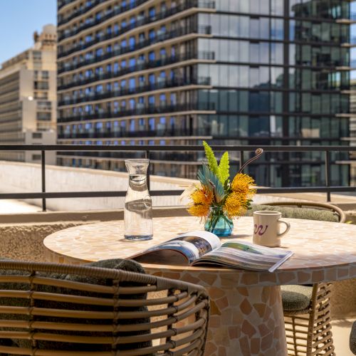 Eine Dachterrasse mit rundem Tisch, St&uuml;hlen, Blumen, einer Zeitschrift, einer Tasse und Geb&auml;uden im Hintergrund in urbaner Umgebung.