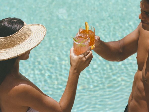 Two people by a pool clink glasses filled with colorful drinks, one wearing a sunhat, enjoying a sunny relaxing moment.
