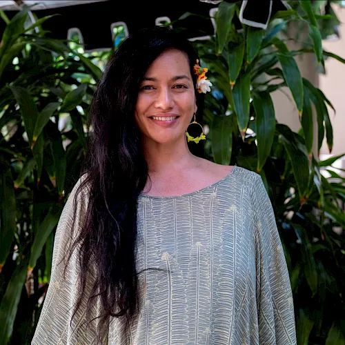 Smiling woman in a light patterned dress with colorful flowers in her hair, standing amid lush green plants.