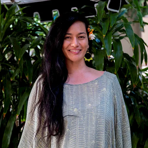 Smiling woman in a light patterned dress with colorful flowers in her hair, standing amid lush green plants.