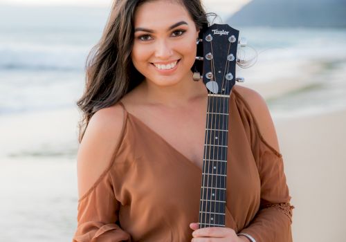 A person stands on a beach holding a guitar, smiling toward the camera, with waves and a mountain in the background.