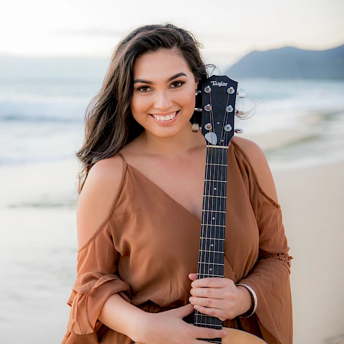 A person stands on a beach holding a guitar, smiling toward the camera, with waves and a mountain in the background.