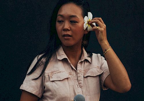 A person in a light outfit holds a microphone and adjusts a flower in their hair against a dark background.