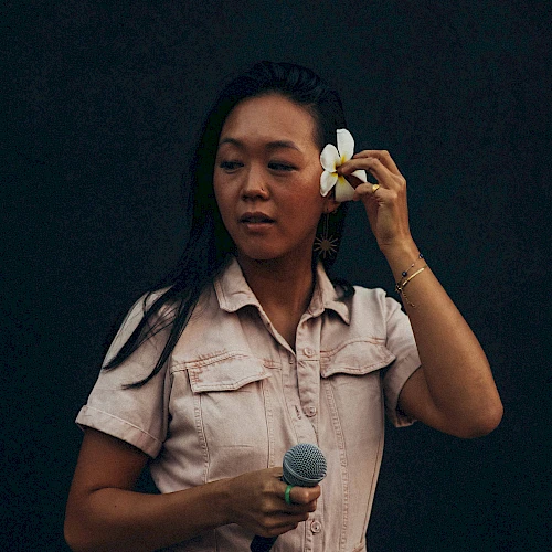 A person in a light outfit holds a microphone and adjusts a flower in their hair against a dark background.