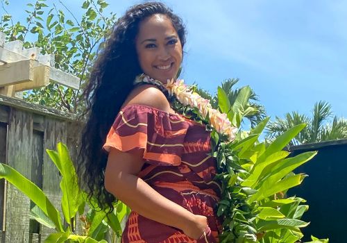 A person in a colorful dress stands outdoors, surrounded by greenery, with a bright sky in the background and a lei around their neck.