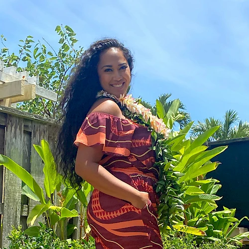 A person in a colorful dress stands outdoors, surrounded by greenery, with a bright sky in the background and a lei around their neck.