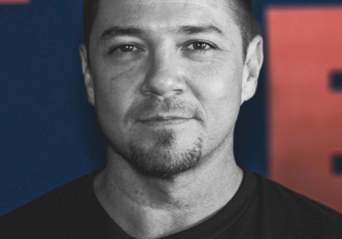The image shows a man with short hair and a goatee, wearing a dark T-shirt, against a background with red and blue colors.