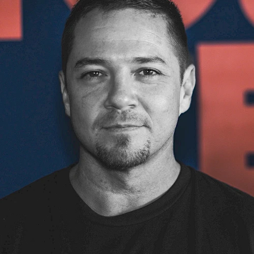 The image shows a man with short hair and a goatee, wearing a dark T-shirt, against a background with red and blue colors.