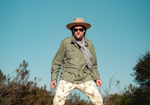 A person stands on a path outdoors, wearing a hat, sunglasses, scarf, green jacket, and patterned pants under a clear blue sky.