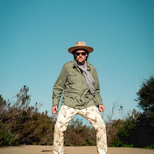 A person stands on a path outdoors, wearing a hat, sunglasses, scarf, green jacket, and patterned pants under a clear blue sky.