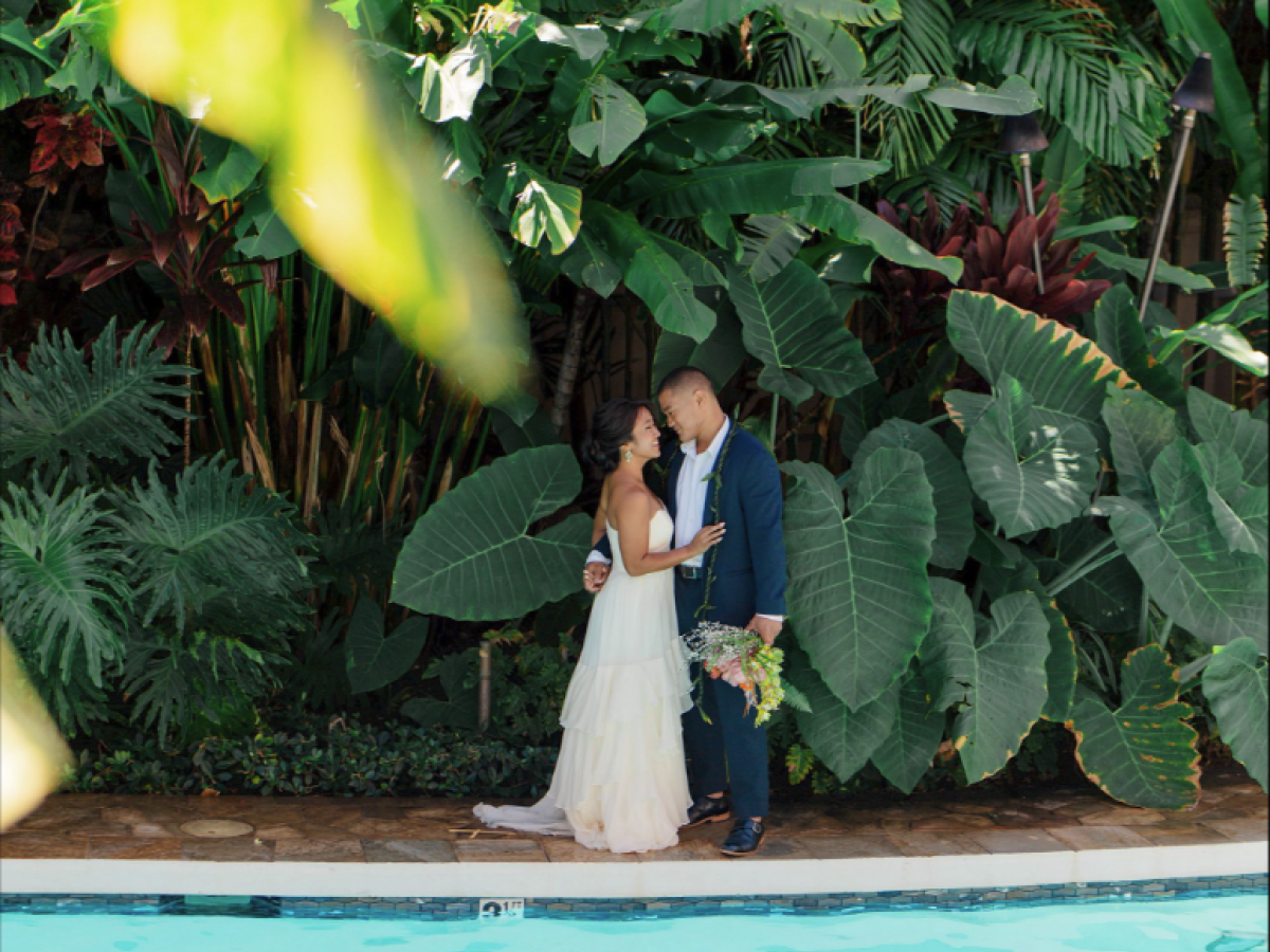 A couple stands by a pool, surrounded by lush green foliage, sharing a romantic moment in the sunlight.