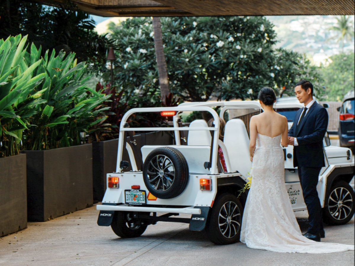A couple in wedding attire stands beside a small white vehicle under a sign reading "Wayfinder," surrounded by lush greenery.