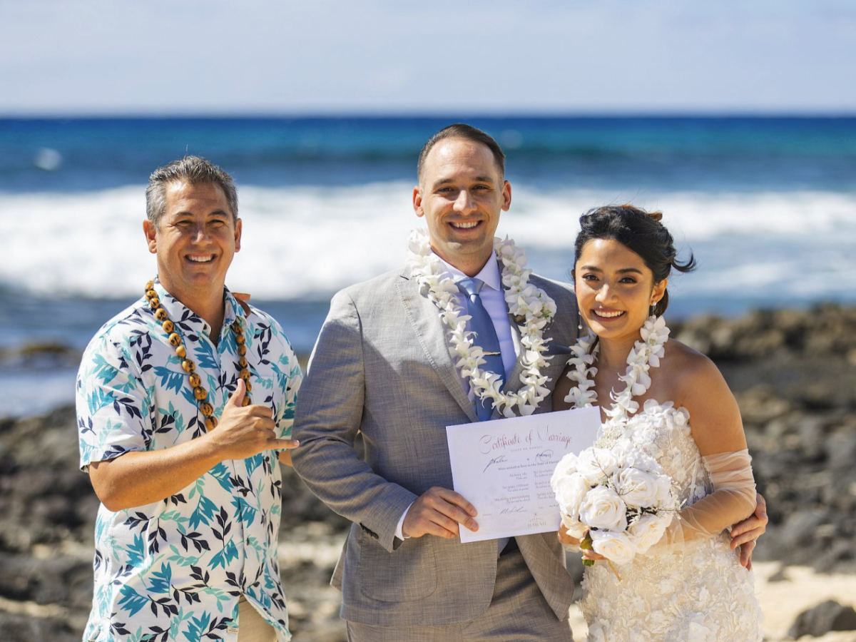 A couple poses with an officiant on a beach; they hold a document and wear leis, celebrating a special occasion.