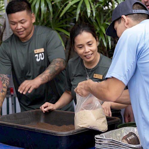 People are gathered around a table with containers, engaging in an activity involving a plastic bag and a tray.