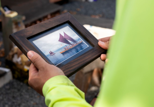 A person in a yellow sleeve holds a framed photo of a sailboat with red sails on the water.