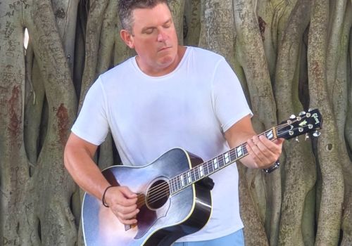 A person is playing a guitar in front of a large tree with exposed roots, which have various engravings on them.