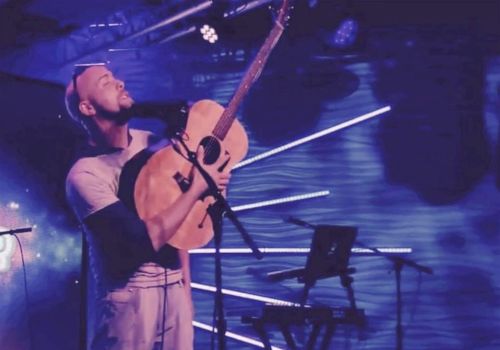 A musician performs passionately with an acoustic guitar on stage, surrounded by vibrant blue lighting and a keyboard stand.