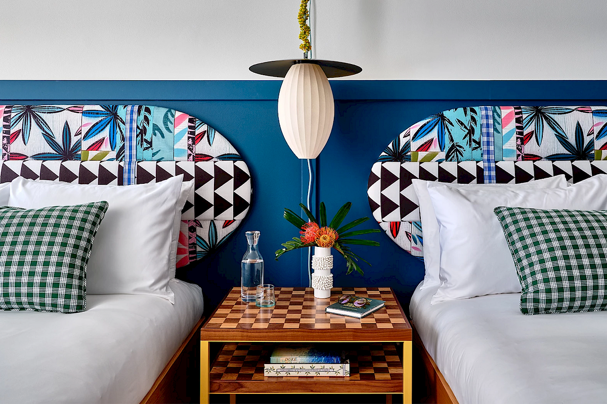 A colorful bedroom with two beds, checkered pillows, a geometric headboard, and a central nightstand with a lamp, books, and flowers.
