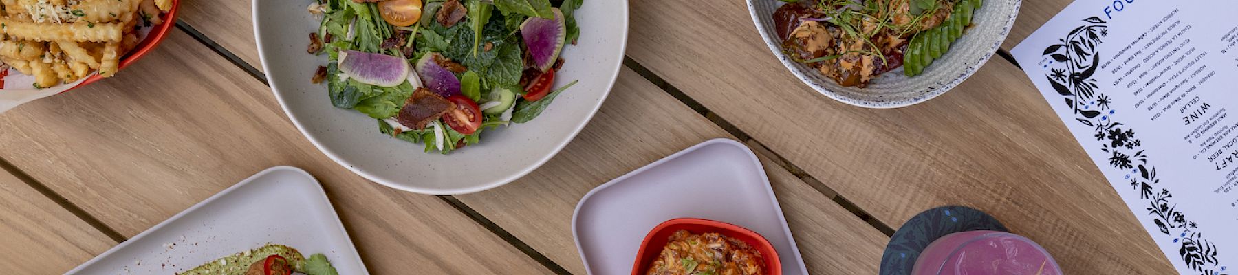 An assortment of dishes on a table, including salads, fries, skewers, and drinks, alongside a visible menu on the top right.