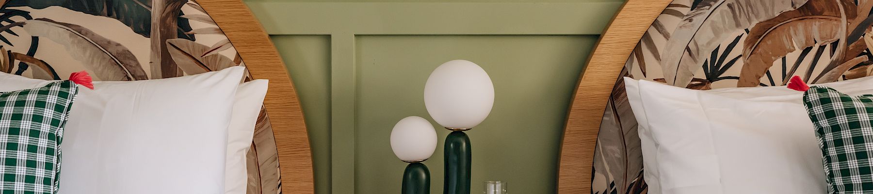 Two twin beds with patterned headboards, green accent wall, checked pillows. A cactus-shaped lamp and books are on a shared bedside table.