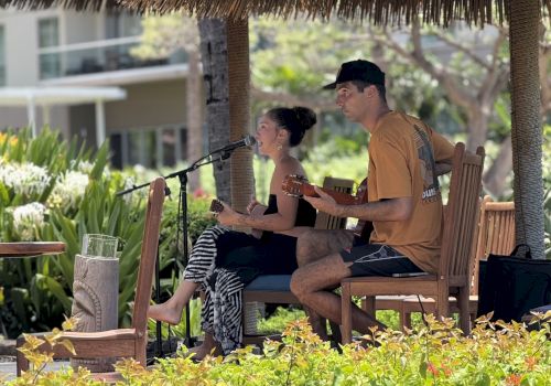 A man and woman are playing guitars and singing under a thatched roof gazebo surrounded by greenery.