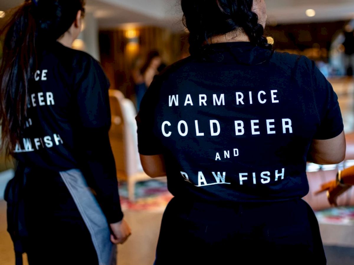 Two individuals are wearing black shirts reading "WARM RICE COLD BEER and RAW FISH" in a restaurant setting.