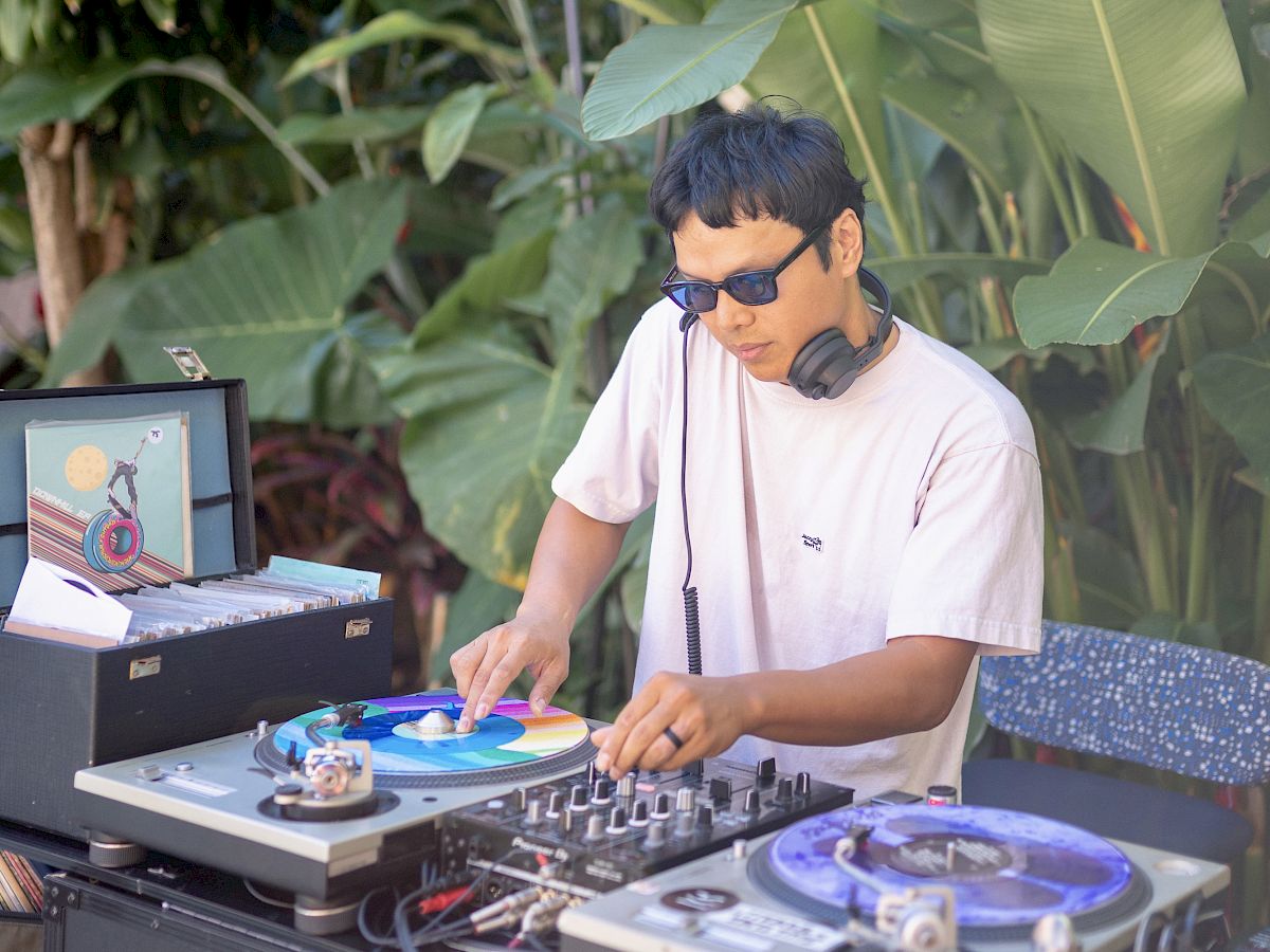 A DJ wearing sunglasses spins on turntables outdoors, surrounded by tropical plants and equipment, mixing tracks with headphones on.