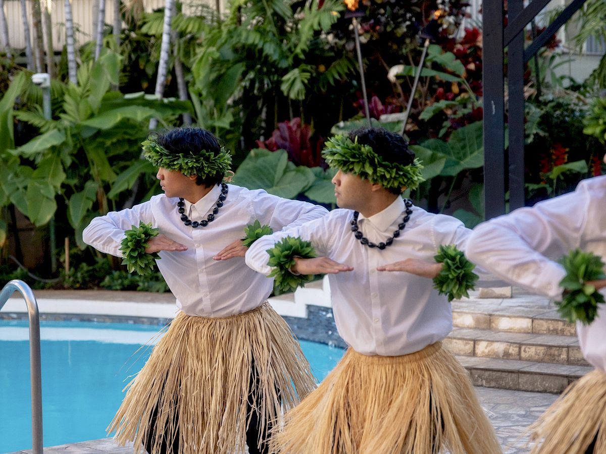 Three people wearing grass skirts and leis perform a Polynesian hula dance by a pool, holding green bouquets in a tropical outdoor setting.