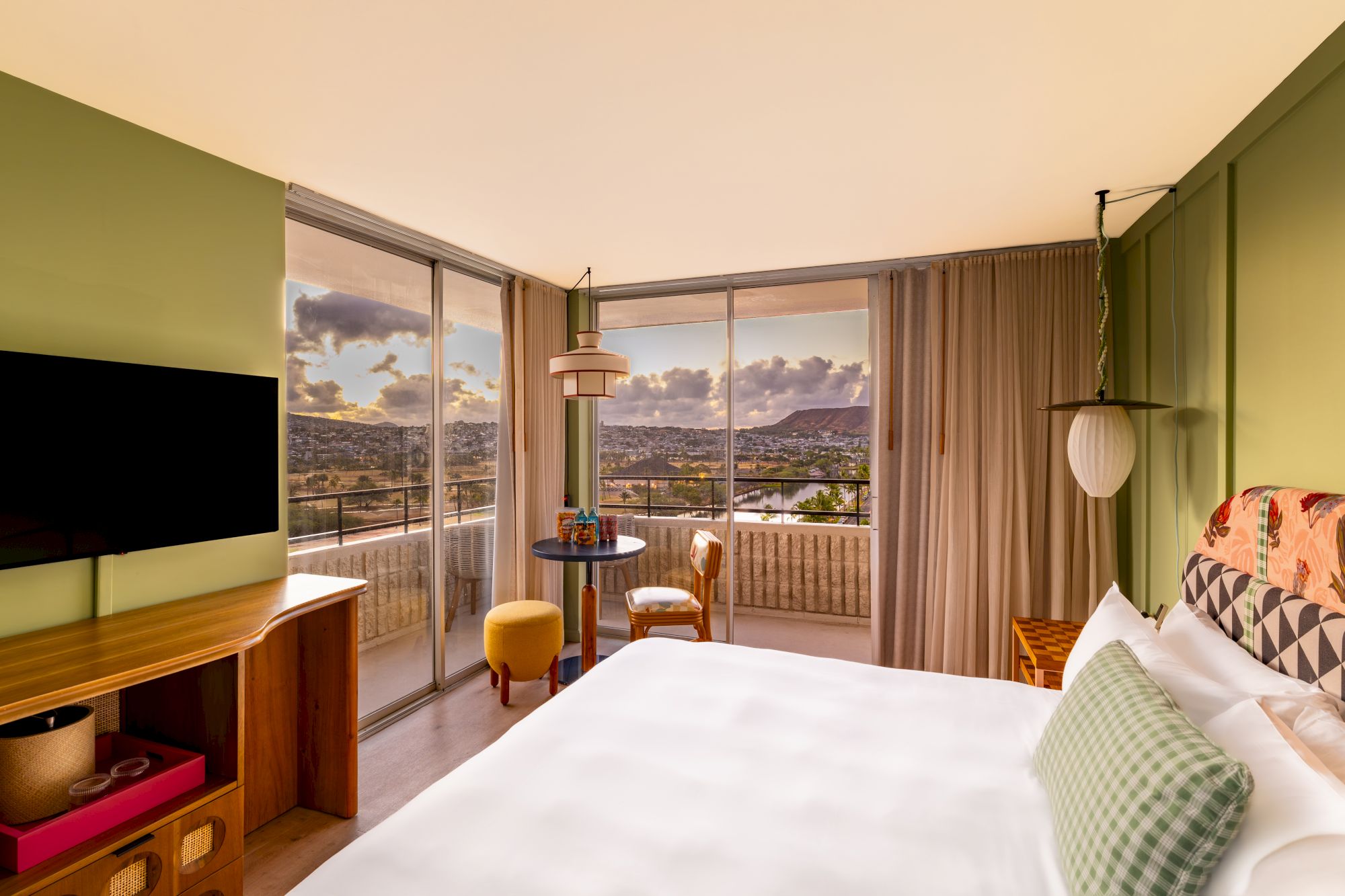A modern hotel room with a large bed, green accent walls, a mounted TV, a small desk, a balcony with city views, and a cozy seating corner by the window.