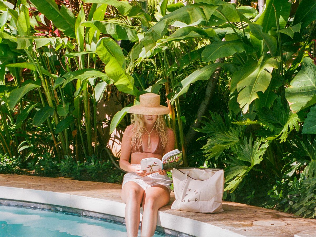 A woman sits poolside by lush tropical plants, wearing a sun hat and reading a book beside a beige tote bag.