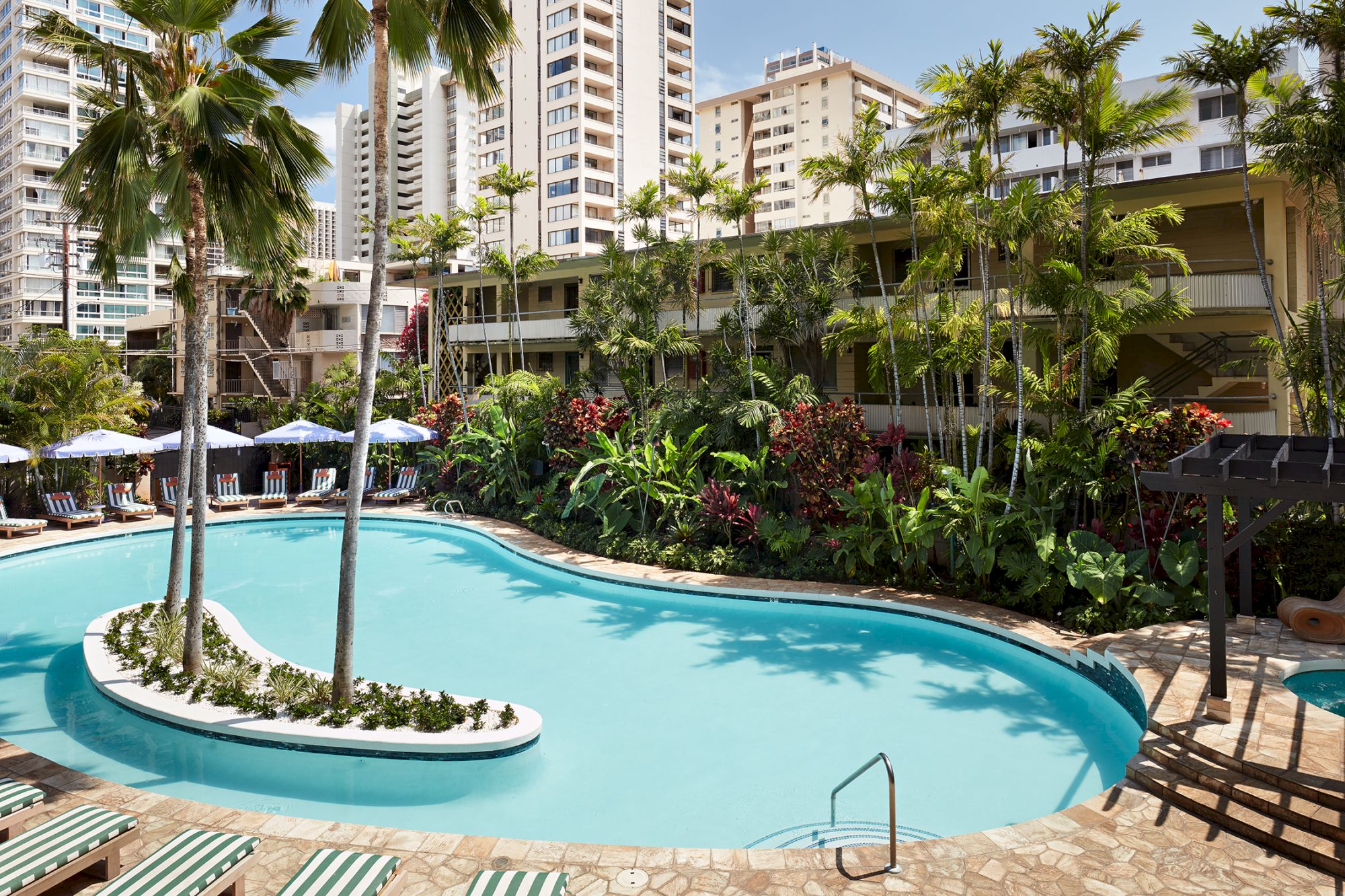 A resort-style pool area with curved blue water, lounge chairs, palm trees, tropical plants, and several mid-rise buildings in the background.