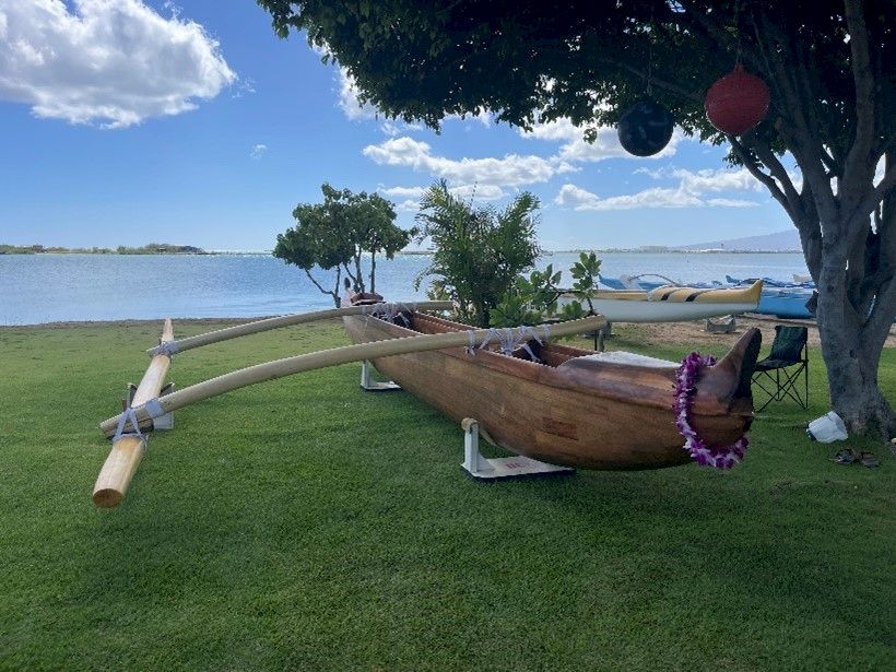 A traditional wooden outrigger canoe on a grassy lakeside, anchored near a tree with hanging decorations and a calm blue water backdrop.