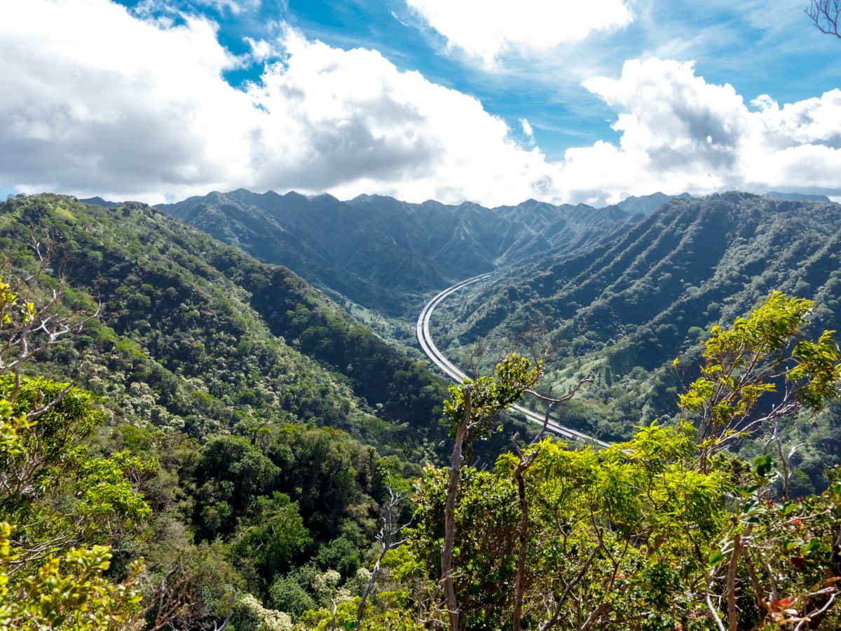 A winding highway cuts through green, mountainous terrain under a bright, partly cloudy sky.