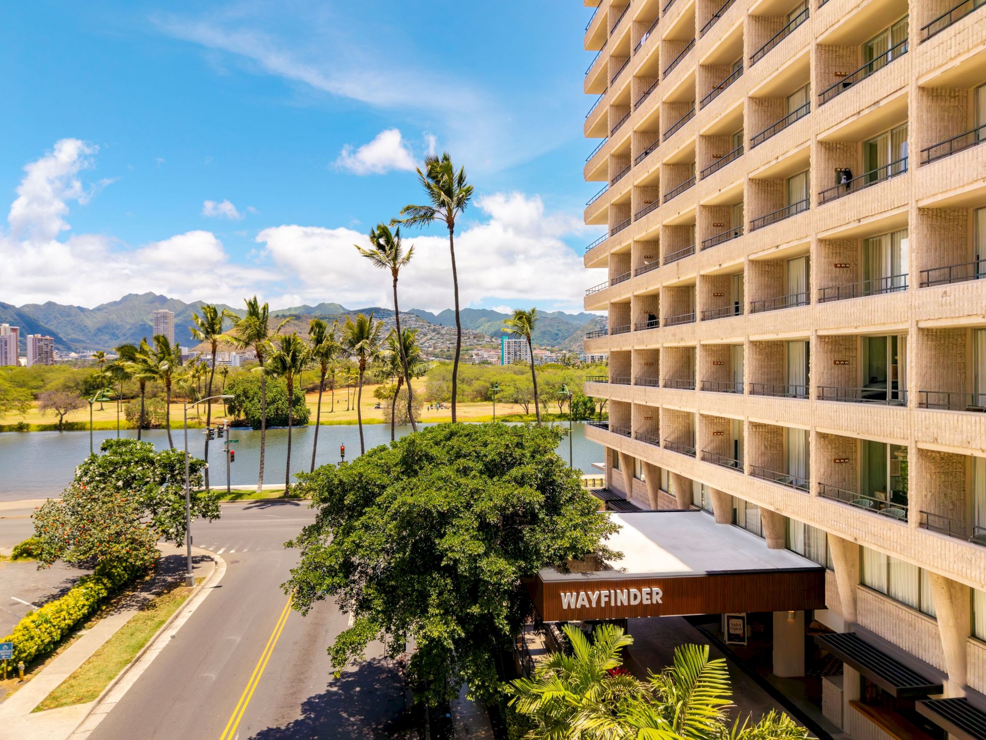 A sunny coastal scene with a tall beige hotel on the right, palm trees, a road, and a sign reading &ldquo;WAYFINDER&rdquo; at the entrance.