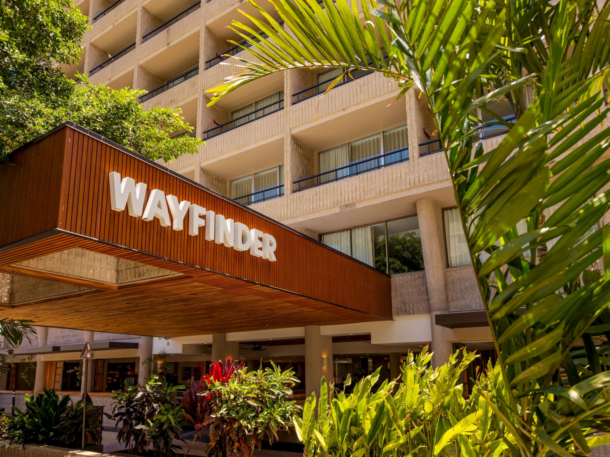 A Wayfinder hotel or building entrance with a wooden sign &ldquo;WAYFINDER,&rdquo; tropical plants, and a multi-story beige facade.
