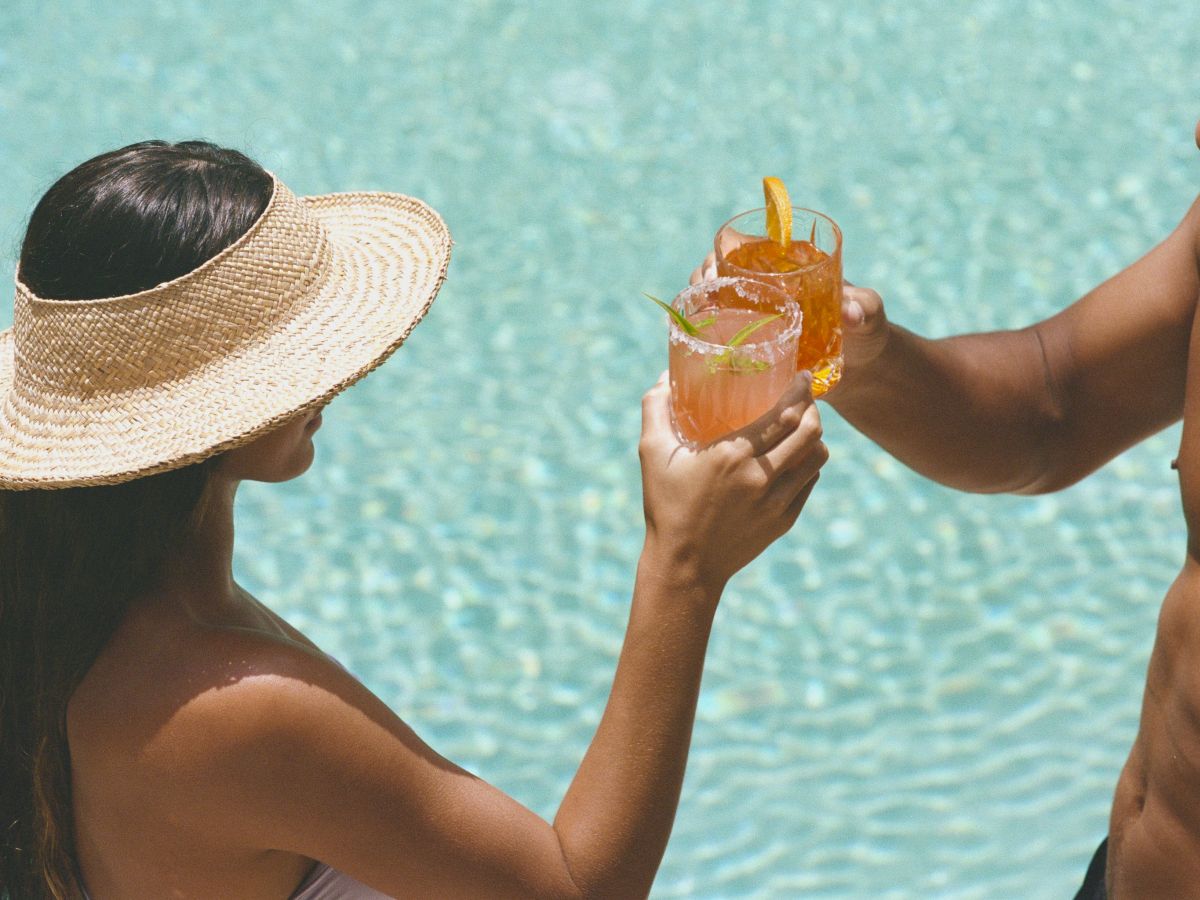 Two people by a pool clinking cocktails; a woman in a sunhat and a man toast with drinks over a blue-tiled pool.