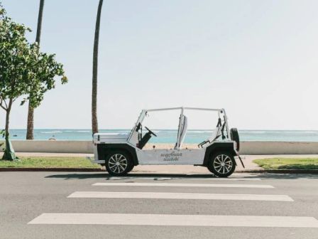 An open-top vehicle is parked beside a beach, with palm trees and the ocean in the background, near a pedestrian crosswalk.