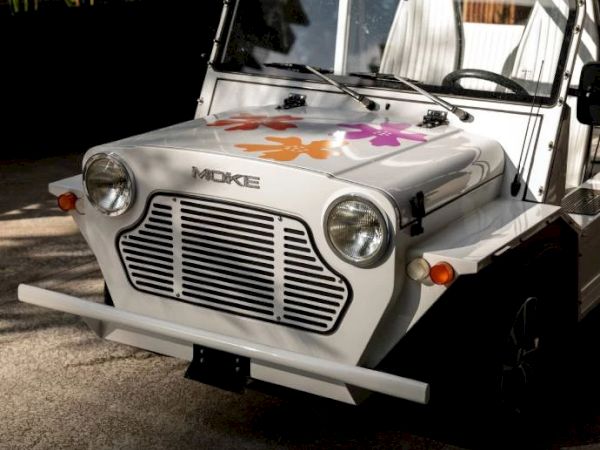 A white MOKE vehicle with a front floral decoration, headlights, and a distinctive grille, parked outdoors.