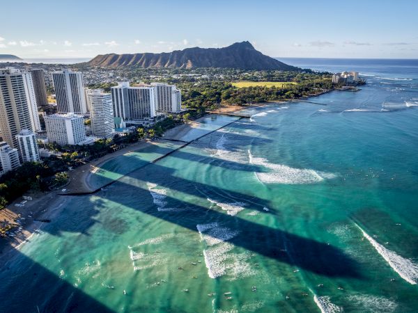 Aerial view of a sunny coastline with high-rise hotels along a sandy beach, turquoise water, and a mountain in the distance, casting long shadows.