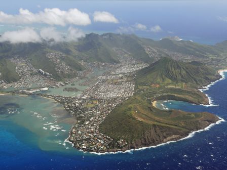 Aerial view of a coastal volcanic landscape with a curving bay, lush green hills, and a town along the shoreline, meeting blue ocean.
