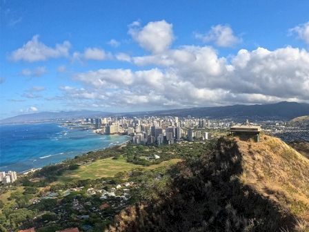A scenic view of a coastal city with a large grassy area, buildings, and shoreline under a partly cloudy sky, with a structure on a hill.
